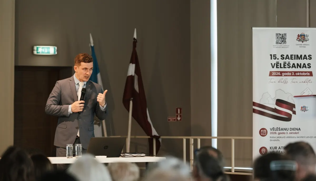Reģionālās apmācības Rīgā — runātājs ar mikrofonu uzrunā dalībniekus; fonā Latvijas karogs un CVK stends ar uzrakstu „15. Saeimas vēlēšanas. 2026. gada 3. oktobris“