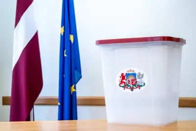 Vēlēšanu kaste ar Latvijas ģerboni uz galda, fonā Latvijas un Eiropas Savienības karogi